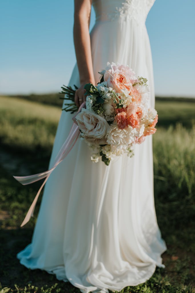 A bride in a flowing white gown holds a colorful bouquet in a sunny field.