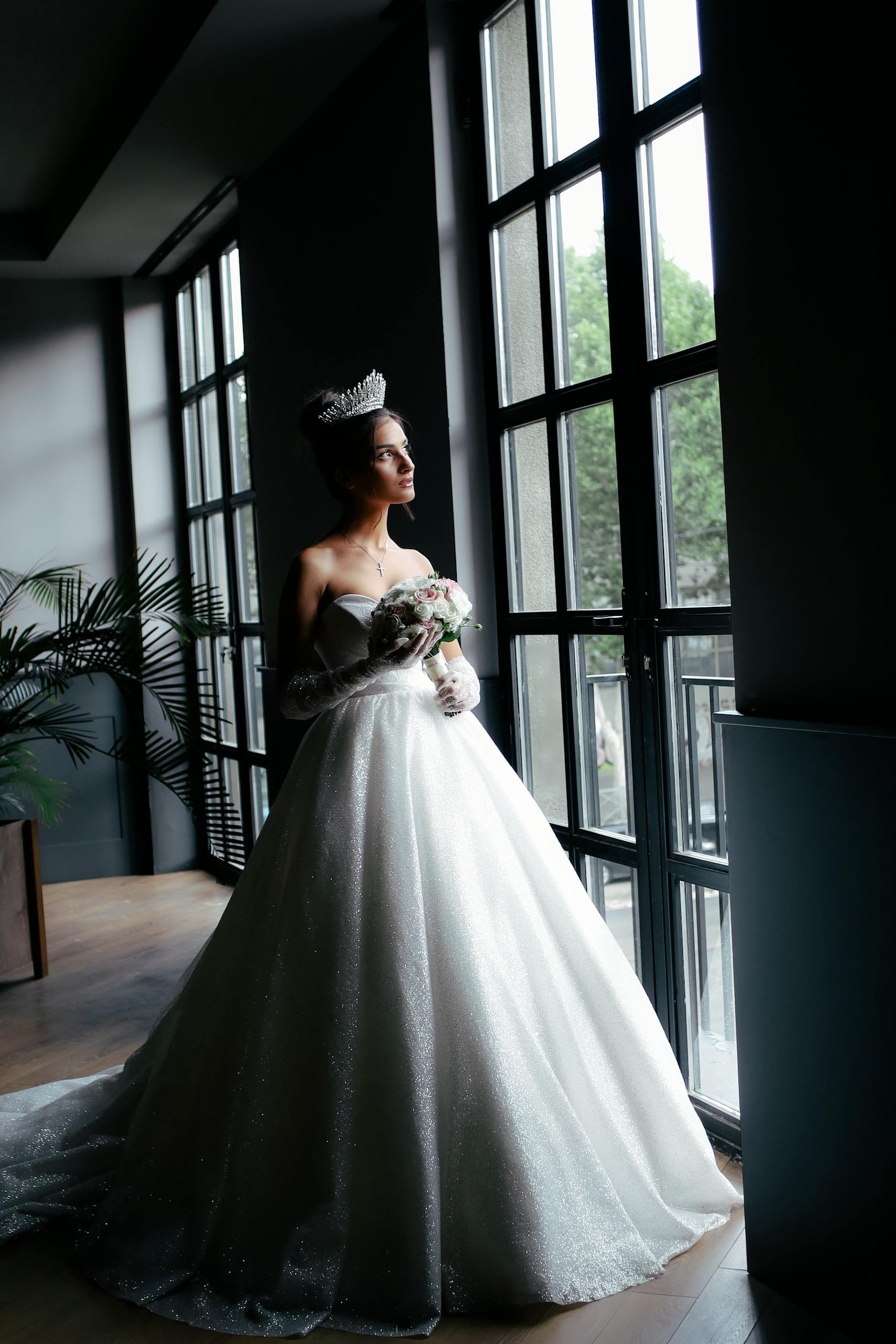 Bride in a sparkling strapless gown holding a bouquet by a large window, wearing a tiara.